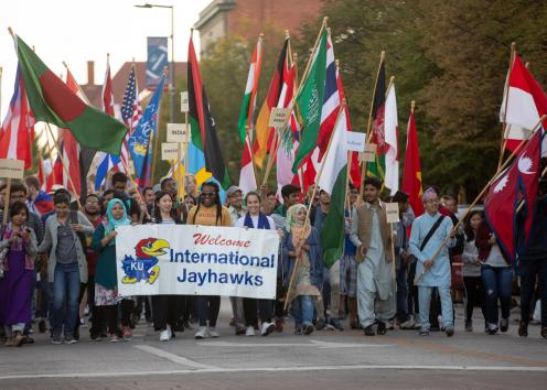 Large group of students walking together hold flags of various nationalities and a Welcome International Jayhawks banner 